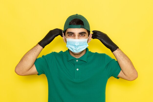 Foto gratuita vista frontal de mensajero de sexo masculino joven en camisa verde gorra verde con máscara estéril sobre el fondo amarillo entregando el color del servicio