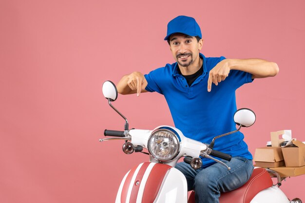 Vista frontal del mensajero sonriente chico con sombrero sentado en scooter y apuntando hacia abajo sobre fondo melocotón pastel