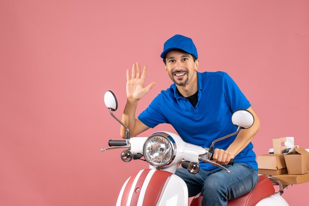 Vista frontal del mensajero sonriente chico con sombrero sentado en scooter mostrando cinco sobre fondo melocotón pastel