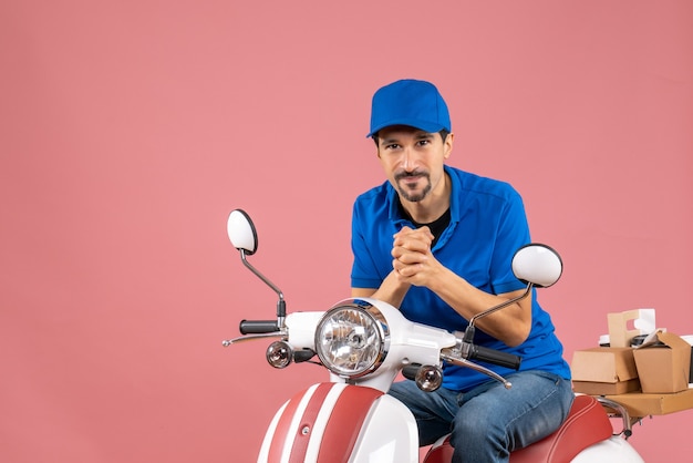 Vista frontal del mensajero sonriente chico con sombrero sentado en scooter sobre fondo de melocotón pastel