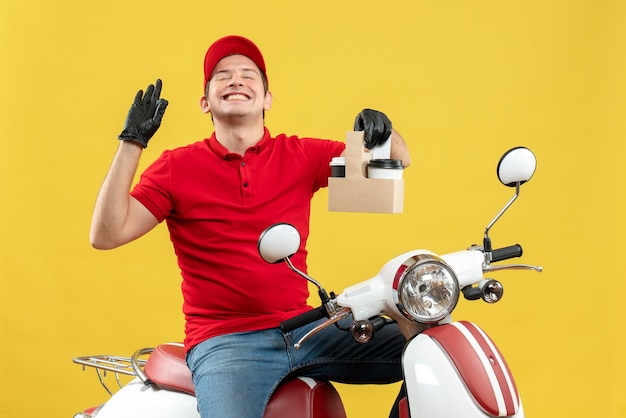 Vista frontal del mensajero sonriente hombre vestido con blusa roja y guantes de sombrero en máscara médica entregando orden sentado en scooter con órdenes