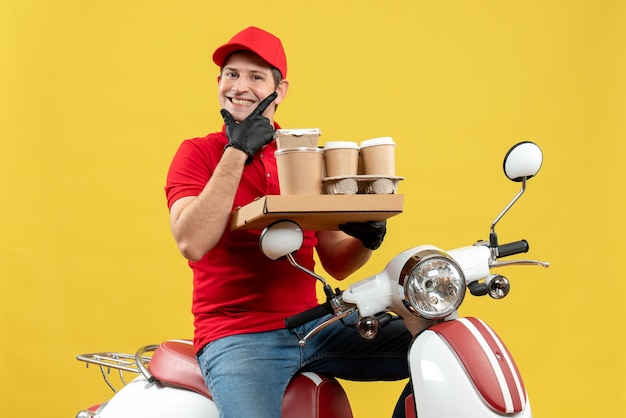 Vista frontal del mensajero sonriente hombre vestido con blusa roja y guantes de sombrero en máscara médica entregando orden sentado en scooter con órdenes