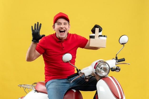 Vista frontal del mensajero sonriente hombre vestido con blusa roja y guantes de sombrero en máscara médica entregando orden sentado en scooter sosteniendo órdenes mostrando cinco
