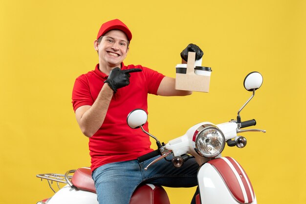 Vista frontal del mensajero sonriente hombre vestido con blusa roja y sombrero guantes en máscara médica entregando orden sentado en scooter señalando órdenes