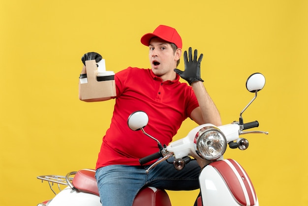 Foto gratuita vista frontal del mensajero sorprendido hombre vestido con blusa roja y guantes de sombrero en máscara médica entregando orden sentado en scooter sosteniendo órdenes mostrando cinco