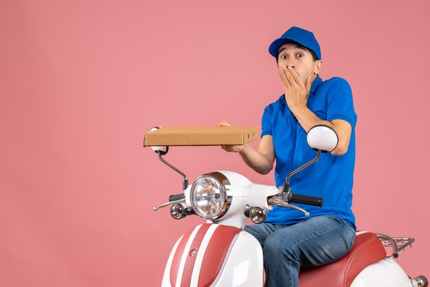 Vista frontal del mensajero sorprendido con sombrero sentado en scooter con orden sobre fondo de melocotón pastel