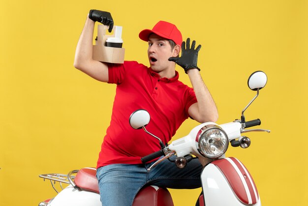 Vista frontal del mensajero sorprendido vestido con blusa roja y guantes de sombrero en máscara médica entregando orden sentado en scooter centrado en pedidos