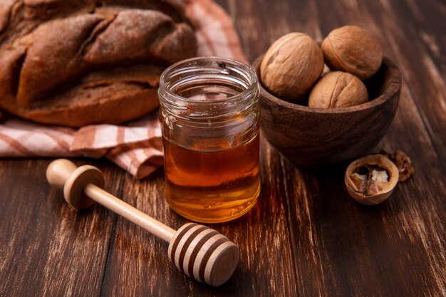 Vista frontal de la miel en un tarro con nueces y una hogaza de pan negro sobre un fondo de madera