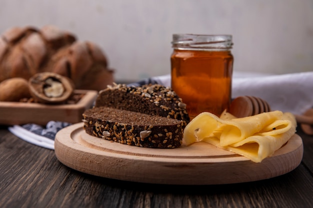 Vista frontal de la miel en un tarro con pan negro y queso sobre un soporte con nueces sobre un fondo de madera