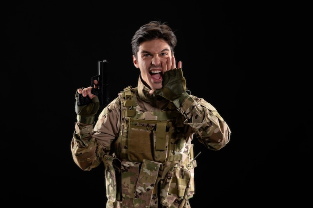 Vista frontal del militar gritando en uniforme con pistola pared negra