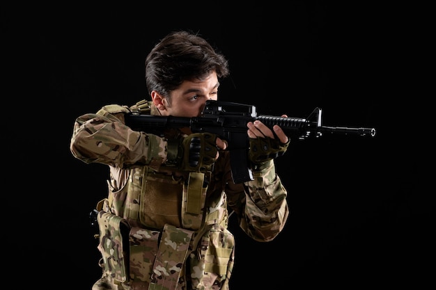 Vista frontal militar militar en uniforme apuntando rifle en pared negra