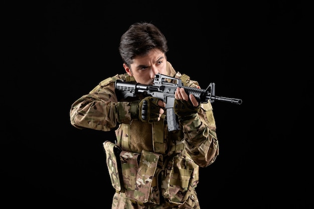 Vista frontal del militar en uniforme apuntando con su rifle de tiro de estudio en la pared negra
