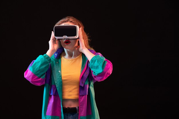 Una vista frontal moderna jovencita con abrigo colorido camiseta naranja jugando realidad virtual en el fondo negro juego interactivo