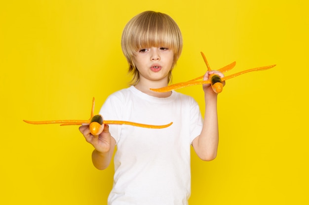 Vista frontal del muchacho rubio con aviones de juguete de color naranja en el escritorio amarillo
