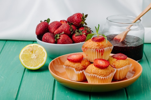 Vista frontal muffins de fresa en un plato con mermelada una rodaja de limón y fresas en un tazón