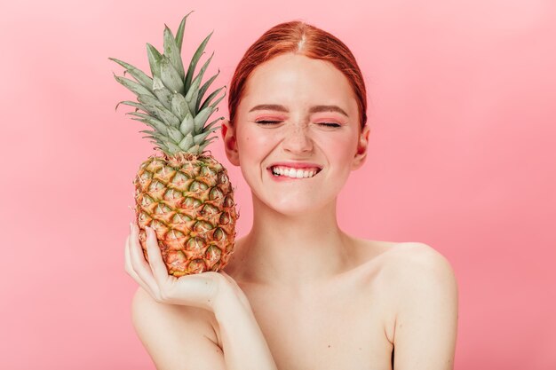 Vista frontal de la mujer alegre con piña posando con los ojos cerrados. Disparo de estudio de chica jengibre emocionada sosteniendo frutas sobre fondo rosa.