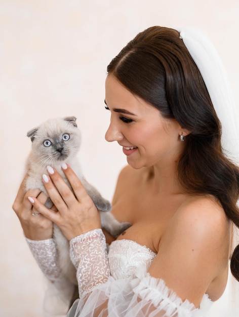 Vista frontal de una mujer atractiva con cabello largo moreno vestido de novia y velo de pie y manteniendo a un gatito doméstico en las manos sonriendo y mirándolo durante el día de la boda