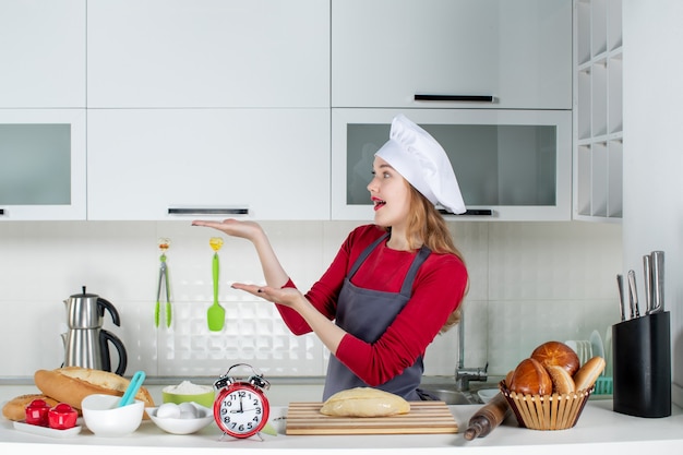 Foto gratuita vista frontal mujer bonita rubia con sombrero de cocinero y delantal de pie detrás de la mesa de la cocina