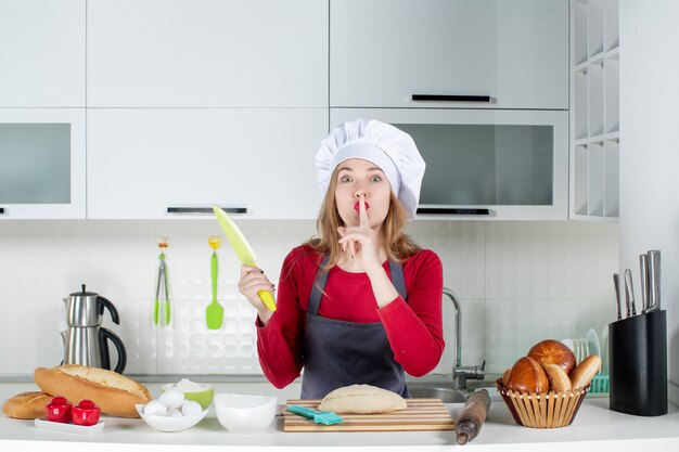 Vista frontal de una mujer bonita con sombrero de cocinero y delantal sosteniendo un cuchillo haciendo letrero de silencio en la cocina