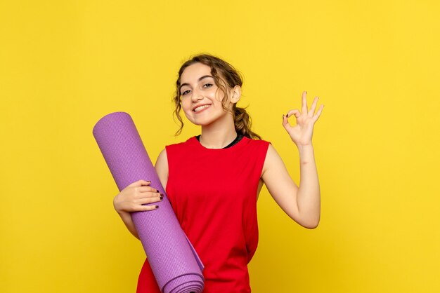 Vista frontal de una mujer bonita sonriendo y sosteniendo una alfombra púrpura sobre amarillo