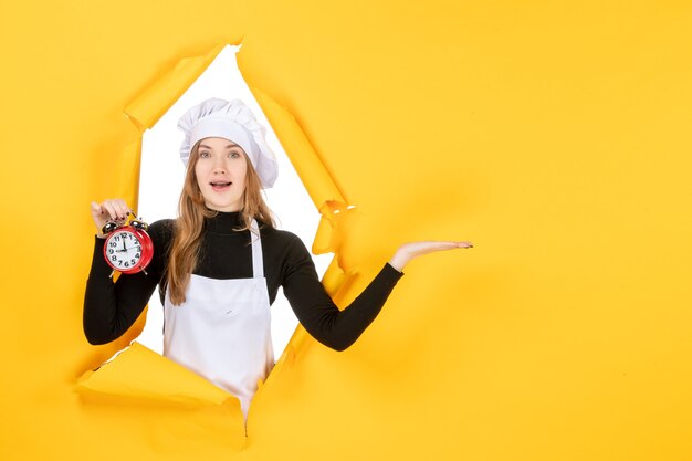 Vista frontal mujer cocinera en gorra de cocinero blanca sosteniendo el reloj en amarillo foto color trabajo emoción cocina cocina sol