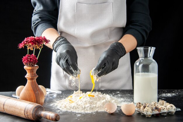 Vista frontal de la mujer cocinera rompiendo los huevos en harina en el trabajo oscuro pastel de pastelería panadería cocinar pastel masa de galletas hornear