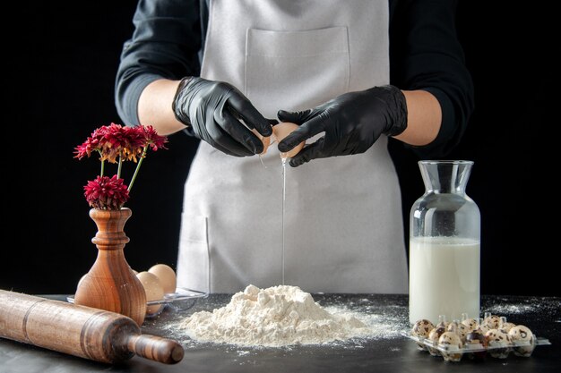 Vista frontal de la mujer cocinera rompiendo los huevos en harina en el trabajo oscuro pastel de pastelería panadería hornear pastel masa de galletas