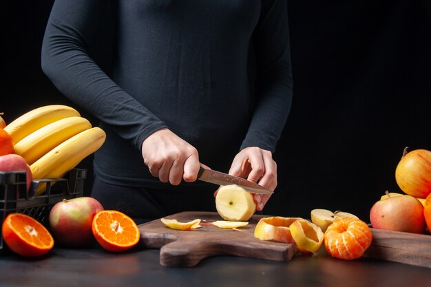 Vista frontal de la mujer cortando manzana fresca sobre tabla de madera frutas en bandeja de madera y caja de plástico en la mesa