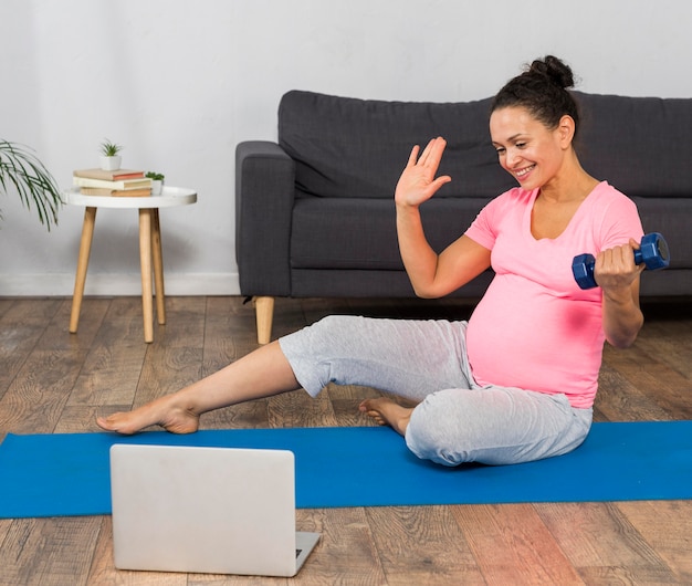 Vista frontal de la mujer embarazada en casa haciendo ejercicio en estera con laptop y peso