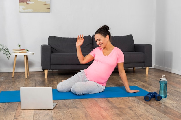 Vista frontal de la mujer embarazada en casa haciendo ejercicio en estera con portátil