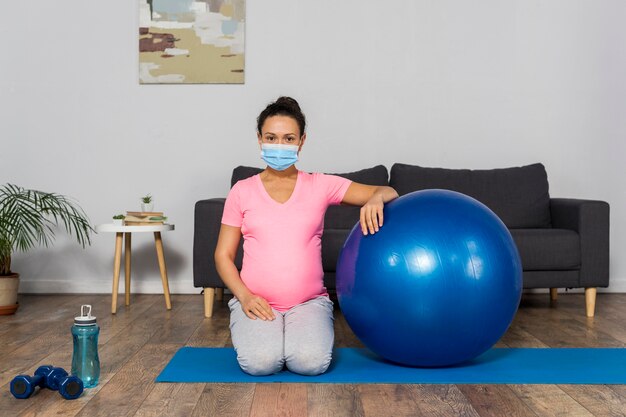 Vista frontal de la mujer embarazada en casa con máscara médica y pelota de ejercicio