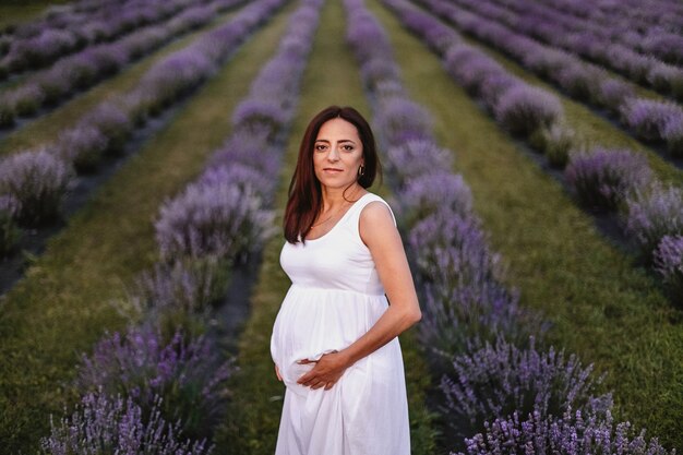 Vista frontal de la mujer embarazada caucásica morena sonriente vestida con un vestido blanco, tocando el vientre en el campo de lavanda.