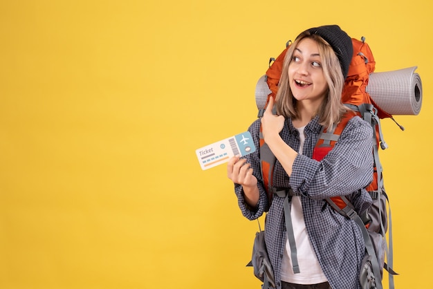 Vista frontal de la mujer feliz viajera con sombrero negro con boleto apuntando a su mochila