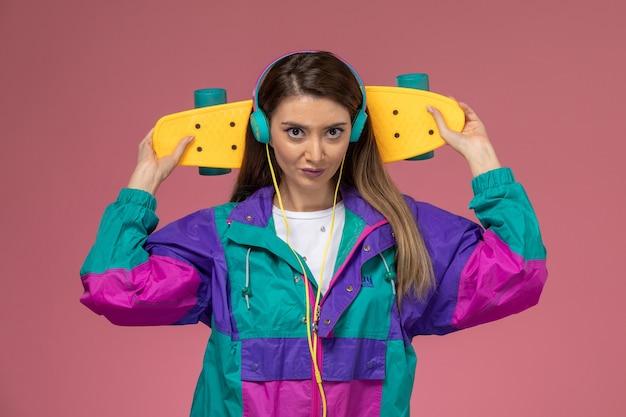 Vista frontal mujer joven con abrigo de color de camisa blanca escuchando música sosteniendo patineta en pared rosa, modelo de pose de mujer de color