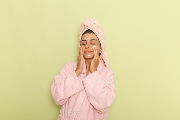 Vista frontal mujer joven en bata de baño rosa aplicando crema facial en un escritorio verde