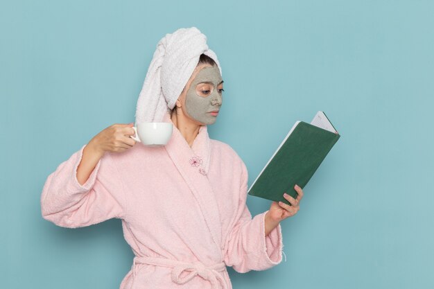 Vista frontal mujer joven en bata de baño rosa bebiendo café y leyendo el cuaderno en el escritorio azul, limpieza, belleza, autocuidado, crema, ducha