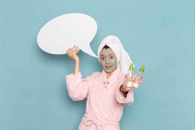 Vista frontal mujer joven en bata de baño rosa con cartel blanco y aerosoles en la pared azul ducha de cuidado personal crema de baño de agua de belleza