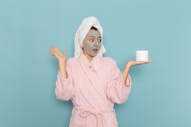 Vista frontal mujer joven en bata de baño rosa con crema para la cara en la pared azul ducha limpieza belleza crema de autocuidado