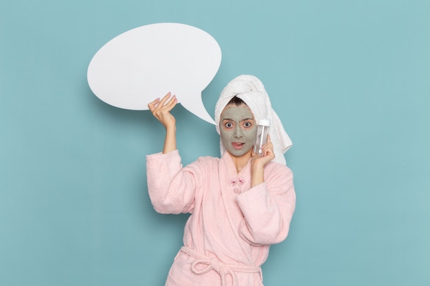 Vista frontal mujer joven en bata de baño rosa después de la ducha con cartel blanco y spray en la pared azul ducha de autocuidado de crema de agua de belleza