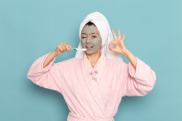 Vista frontal mujer joven en bata de baño rosa después de la ducha limpiando sus dientes en la pared azul claro belleza agua autocuidado ducha limpia