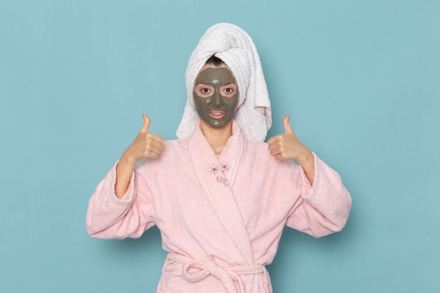 Vista frontal mujer joven en bata de baño rosa después de la ducha con máscara oscura en su rostro en el escritorio azul belleza agua crema autocuidado ducha baño