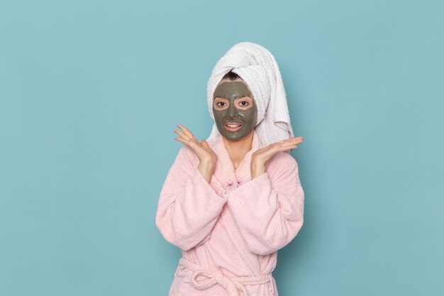 Vista frontal mujer joven en bata de baño rosa después de la ducha con máscara sonriendo en la pared azul belleza agua crema autocuidado ducha baño