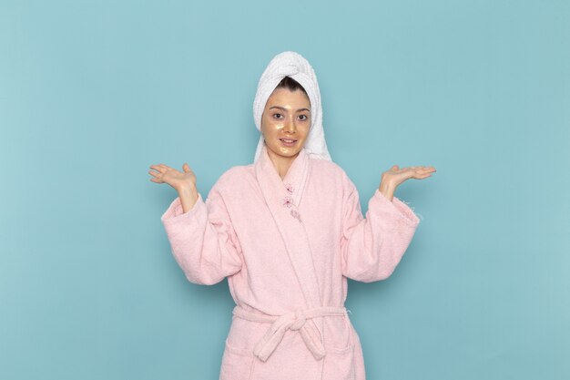 Vista frontal mujer joven en bata de baño rosa después de la ducha sonriendo en la pared azul ducha de autocuidado de crema de agua de belleza