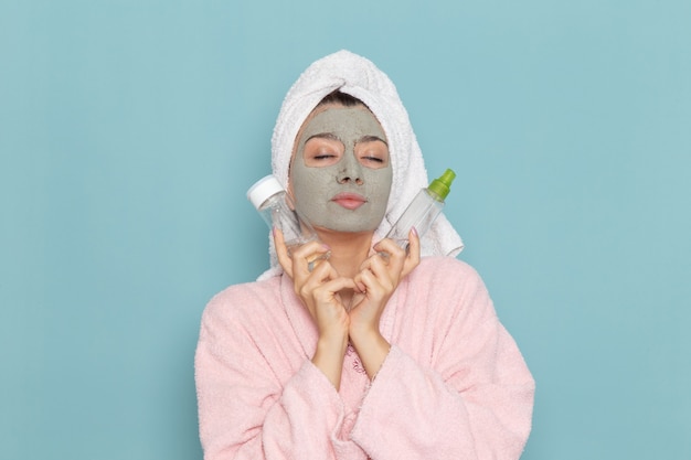 Vista frontal mujer joven en bata de baño rosa después de la ducha sosteniendo aerosoles en la pared azul claro belleza agua crema autocuidado ducha baño