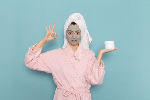 Vista frontal mujer joven en bata de baño rosa con máscara en la cara con crema en el escritorio azul ducha limpieza belleza crema de autocuidado