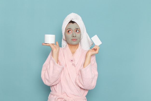 Vista frontal mujer joven en bata de baño rosa con máscara en su rostro sosteniendo la tarjeta en la pared azul ducha limpieza belleza crema de cuidado personal