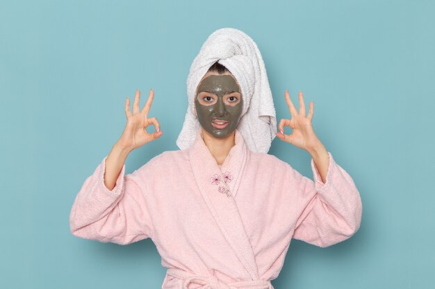 Vista frontal mujer joven en bata de baño rosa posando con máscara en su rostro en la pared azul ducha de autocuidado de crema de baño de agua de belleza