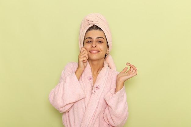 Vista frontal mujer joven en bata de baño rosa posando y sonriendo en un escritorio verde