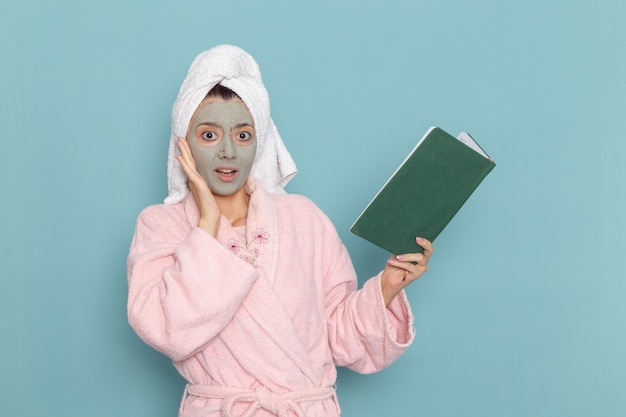 Vista frontal mujer joven en bata de baño rosa sosteniendo y leyendo el cuaderno en la pared azul ducha de autocuidado de crema de baño de agua de belleza