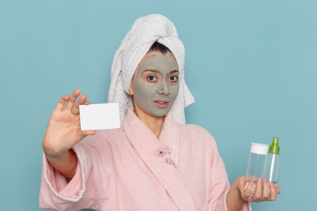 Vista frontal mujer joven en bata de baño rosa con tarjeta y aerosoles en la pared azul ducha de autocuidado de crema de baño de agua de belleza